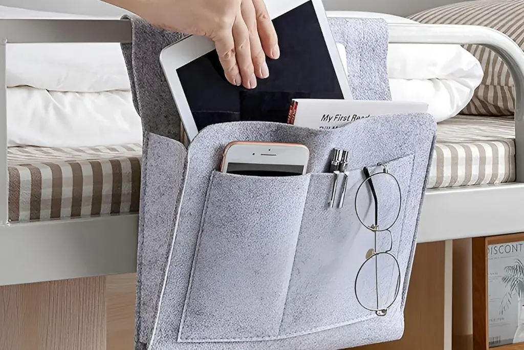 Bedside pocket organizer hanging from a bed, holding a book, phone and glasses.