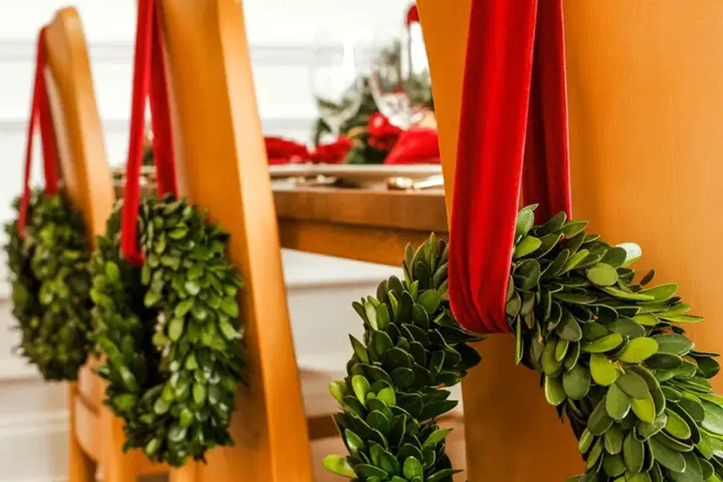 Small green wreaths with red ribbons hanging on the backs of wooden chairs.