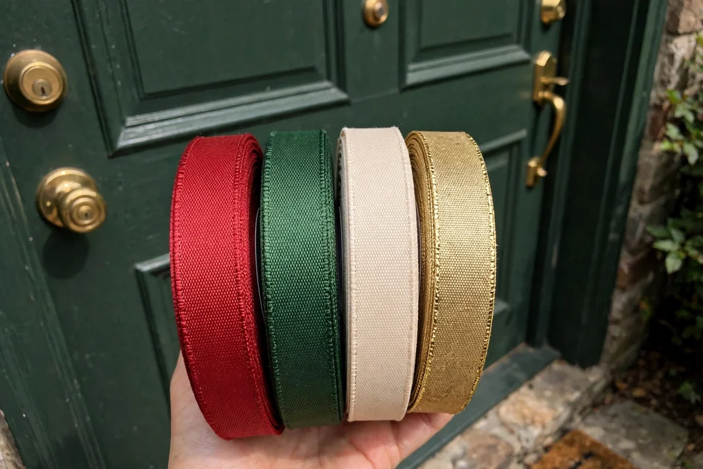 Rolls of red, green, cream, and gold wired ribbon held against a dark green front door in daylight.