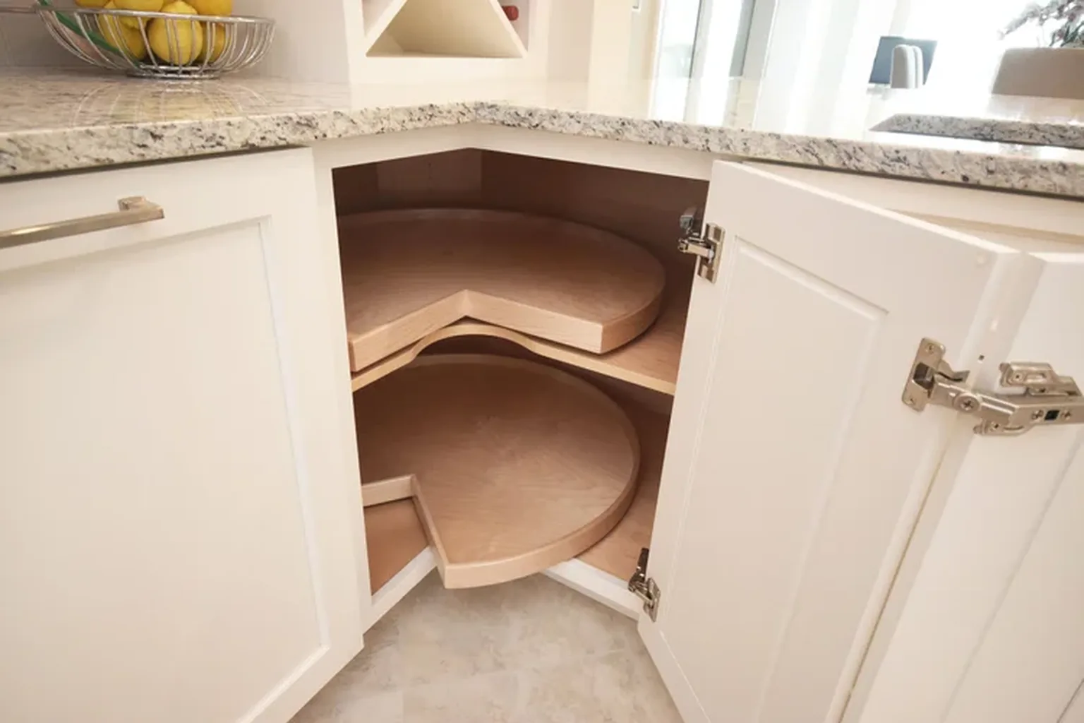 Open white corner base cabinet with wooden lazy Susan shelves under a granite counter, bowl of lemons nearby.