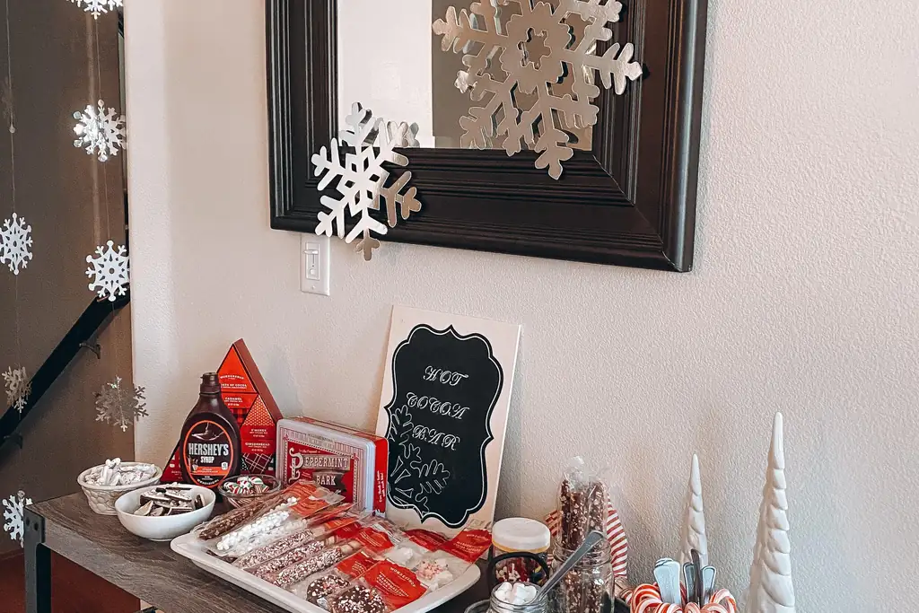Table against a wall with a cocoa tray, toppings, bottles, and a small chalkboard sign.