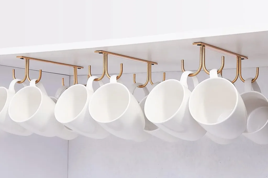 White mugs hanging from gold hooks on a rail under a cabinet.