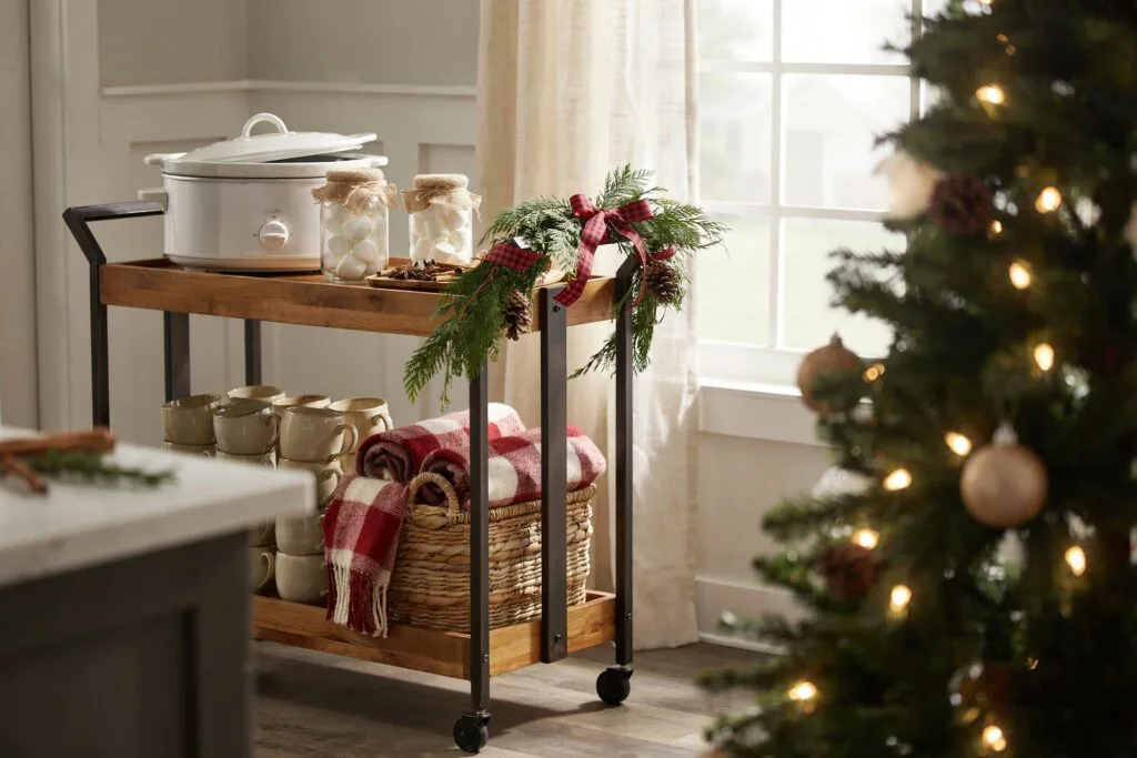 Two tier rolling cart with jars, mugs, and a basket set up as a cocoa bar beside a Christmas tree.
