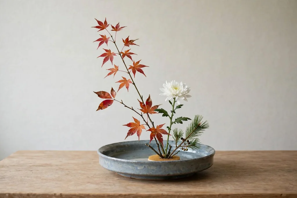 Simple ikebana arrangement with a few seasonal stems in a shallow ceramic dish on a wood table.
