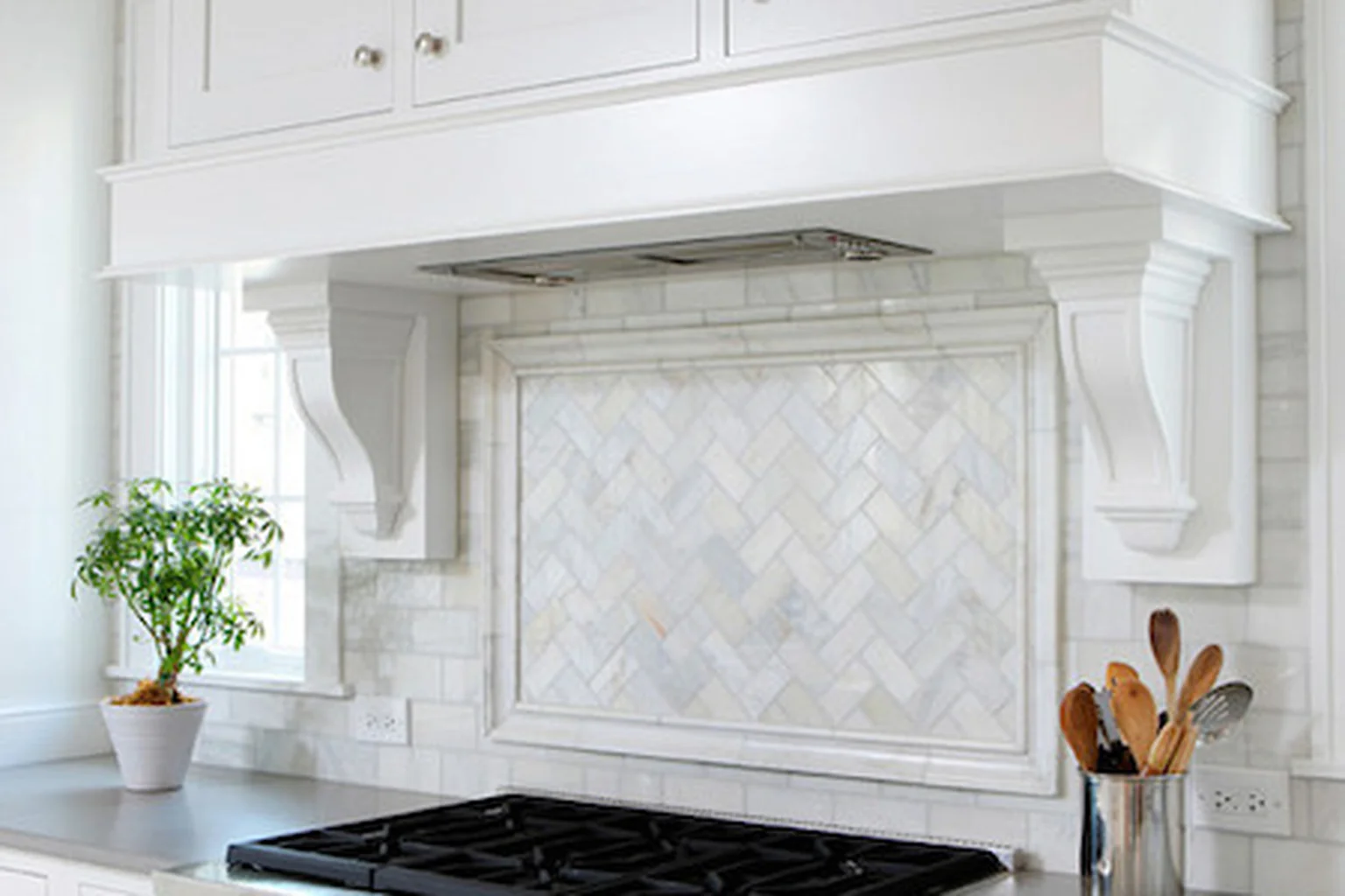 White painted range hood surround over a stove with a marble herringbone backsplash.