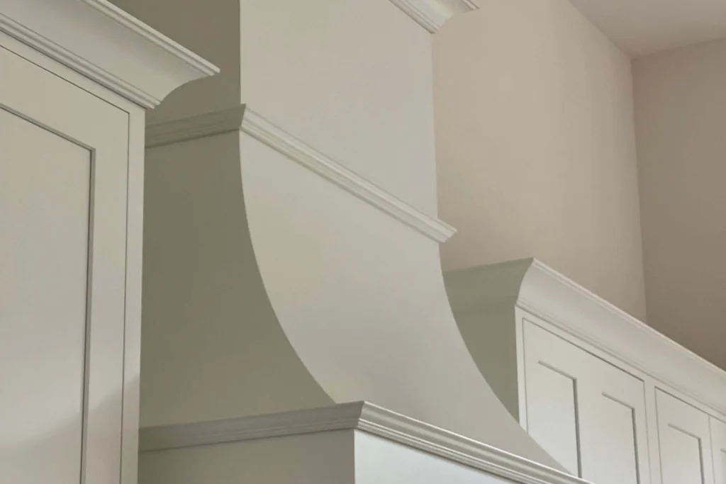 White painted chimney range hood with simple trim fitted between white kitchen cabinets.