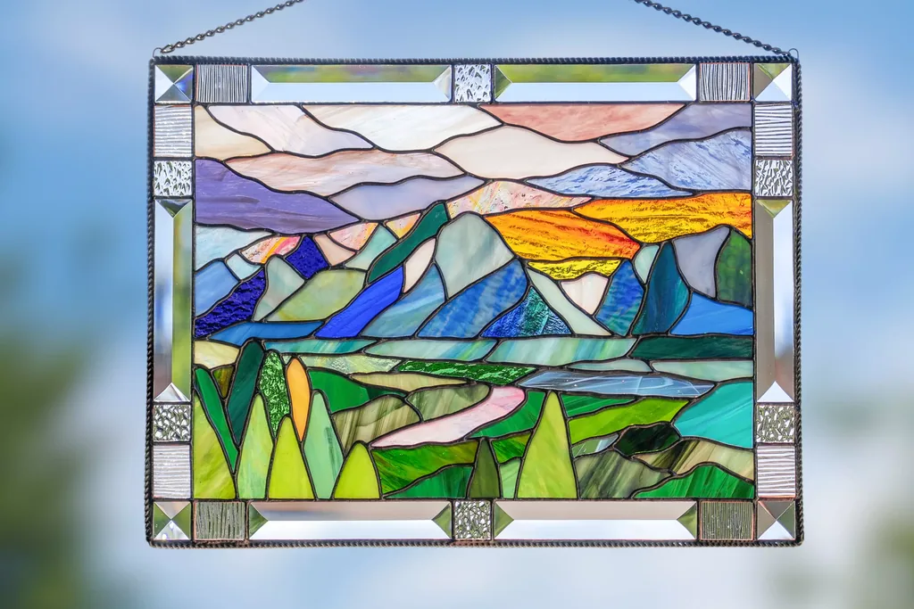 Stained glass panel with a colored landscape design hanging by chains in front of a window.