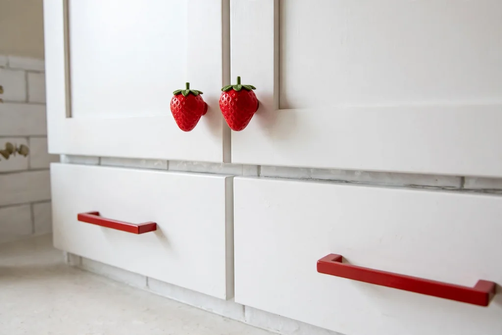 White kitchen cabinets with strawberry shaped knobs and red drawer pulls.