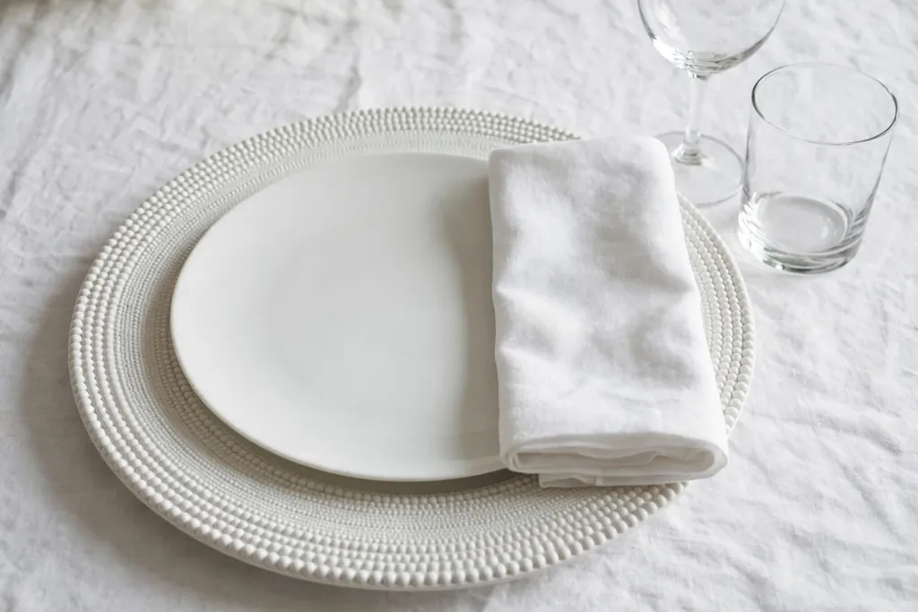 White place setting with a textured white charger, plain white plate, and folded napkin.
